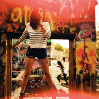 A woman wearing a white long-sleeve striped crop top and black denim shorts stands in front of a graffiti-covered wooden frame, with a blurred outdoor street scene behind her.