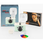 Half Lomovrette Frame instant camera kit with a mint green and cream camera body, a large white flash, its blue retail box, a smaller printed insert card, a round white lens cover, and a small multicolor color card on a white background.