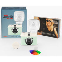 Half Lomovrette Frame instant camera kit with a mint green and cream camera body, a large white flash, its blue retail box, a smaller printed insert card, a round white lens cover, and a small multicolor color card on a white background.