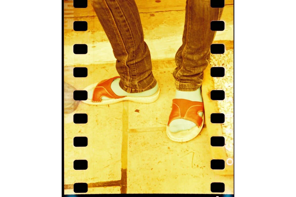 A close-up lifestyle shot of a person's legs in brown jeans wearing orange and white slide sandals, set against a warm yellow-toned tiled floor with a film strip graphic border.