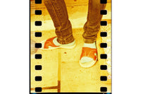 A close-up lifestyle shot of a person's legs in brown jeans wearing orange and white slide sandals, set against a warm yellow-toned tiled floor with a film strip graphic border.