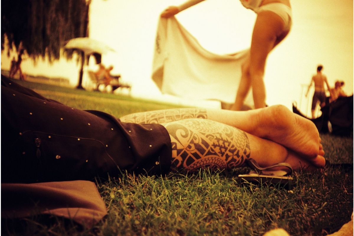 Person lying on grass with tattooed feet, blurred background of people and nature.