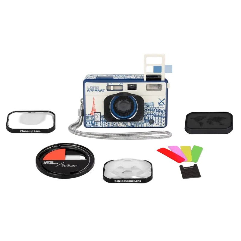A compact camera with a blue and white Paris-themed graphic print, shown with multiple accessories including a close-up lens, a spitzer lens, a kaleidoscope lens, colorful filter pieces, and a black case on a light gray background.