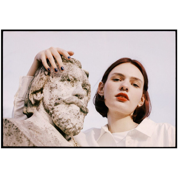 A fashion photograph featuring a woman with short auburn hair, red lipstick, and dark nail polish posing next to a weathered stone bust under a pale sky. She is wearing a white collared shirt.