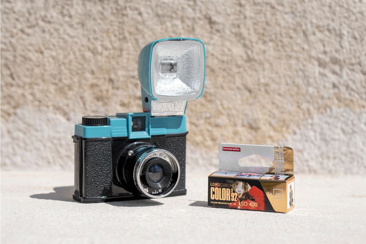 A retro-style instant camera with a turquoise and black body and an attached flash, shown next to a box of Colorplus Color ISO 600 film on a light concrete surface against a textured beige wall.