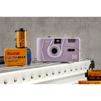 A lavender Kodak Mini 35mm instant camera displayed on a white metal shelf alongside vintage yellow and blue Kodak UltraMax 400 film rolls against a textured gray background.