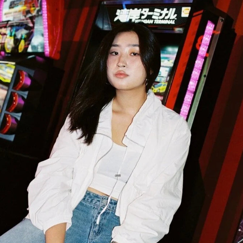 A woman wearing a white oversized jacket over a white crop top and blue jeans, sitting in front of arcade machines in a brightly lit arcade.