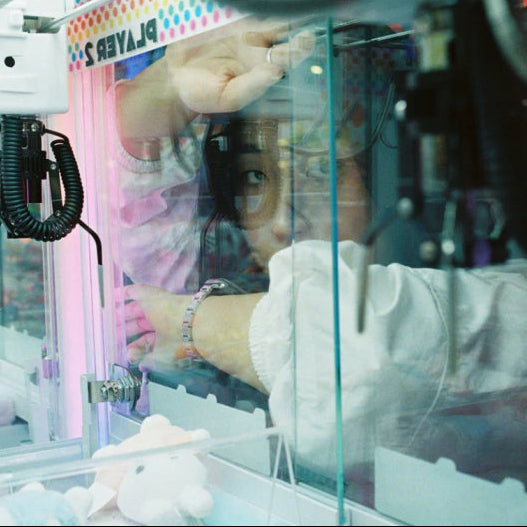 A person in a white shirt reaches inside a claw machine at an arcade, viewed through the glass enclosure with visible reflections and interior lighting.