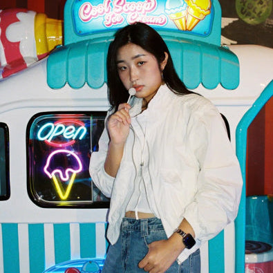 A woman stands in front of a decorative ice cream truck display, holding a small ice cream cone to her mouth. She is wearing a white button-up shirt and blue jeans.