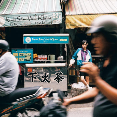 Street scene with people on motorcycles and a person standing in front of a store with signs in an Asian language.