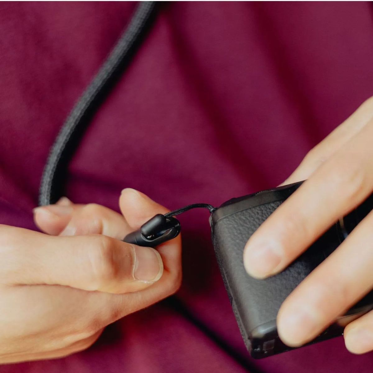 Close-up of a person's hands holding a black textured camera strap or lanyard attachment next to a black camera body, with a burgundy top in the background.