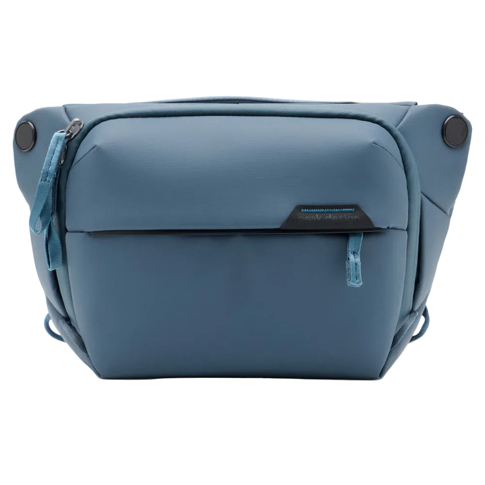 A blue compact camera bag with a front zippered pocket and a small black brand plaque, photographed on a clean white background.