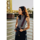 Woman with tattoos holding a camera, standing by a waterfront with a bridge in the background.
