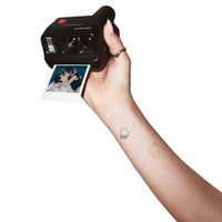 A hand holding a black Polaroid instant camera that is pulling a printed photo strip out, shown against a plain white background.