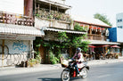 A person riding a black scooter on a street in front of rustic buildings with awnings, hanging red lanterns, and outdoor seating under red umbrellas.