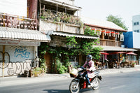 A person riding a black scooter on a street in front of rustic buildings with awnings, hanging red lanterns, and outdoor seating under red umbrellas.