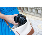 A person in a bright blue dress holds a Canon DSLR camera inside a white and brown shoulder bag with gold-tone hardware, photographed outdoors on a paved walkway.