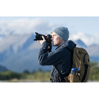 A man wearing a gray beanie, dark jacket, and a brown hiking backpack with a water bottle, holding a camera up to his eye outdoors with mountains in the background.