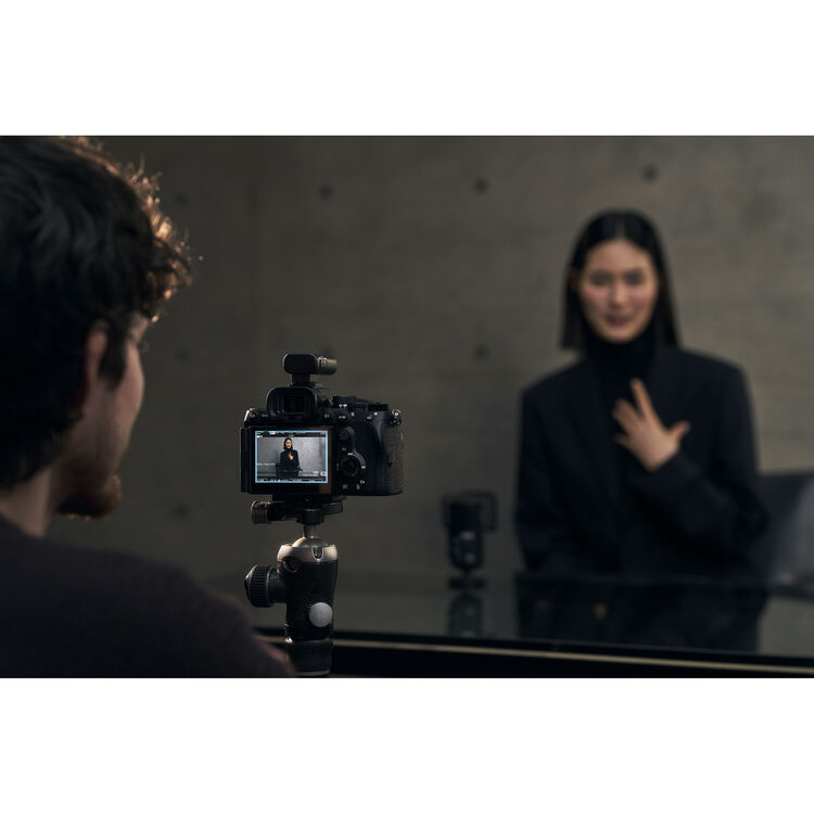 A camera on a tripod records a woman in a black outfit in front of a mirror, with the woman's reflection visible and a blurred figure in the foreground.