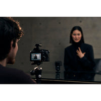 A camera on a tripod records a woman in a black outfit in front of a mirror, with the woman's reflection visible and a blurred figure in the foreground.