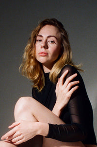 A woman with blonde hair sits cross-legged against a dark gray studio background, wearing a black long-sleeve top with sheer mesh sleeves.