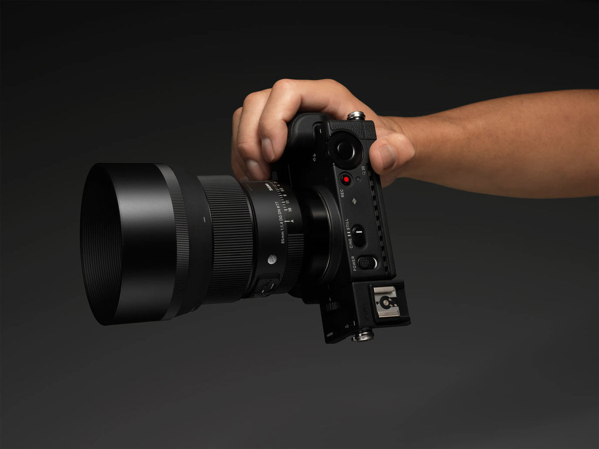 A hand holding a black film camera with a large lens attached, shown against a dark gray studio background.