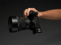 A hand holding a black film camera with a large lens attached, shown against a dark gray studio background.