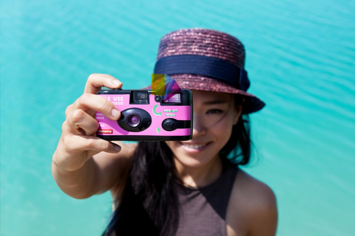 A woman in a pink and navy hat holds a pink disposable camera toward the viewer, with a bright turquoise water background.