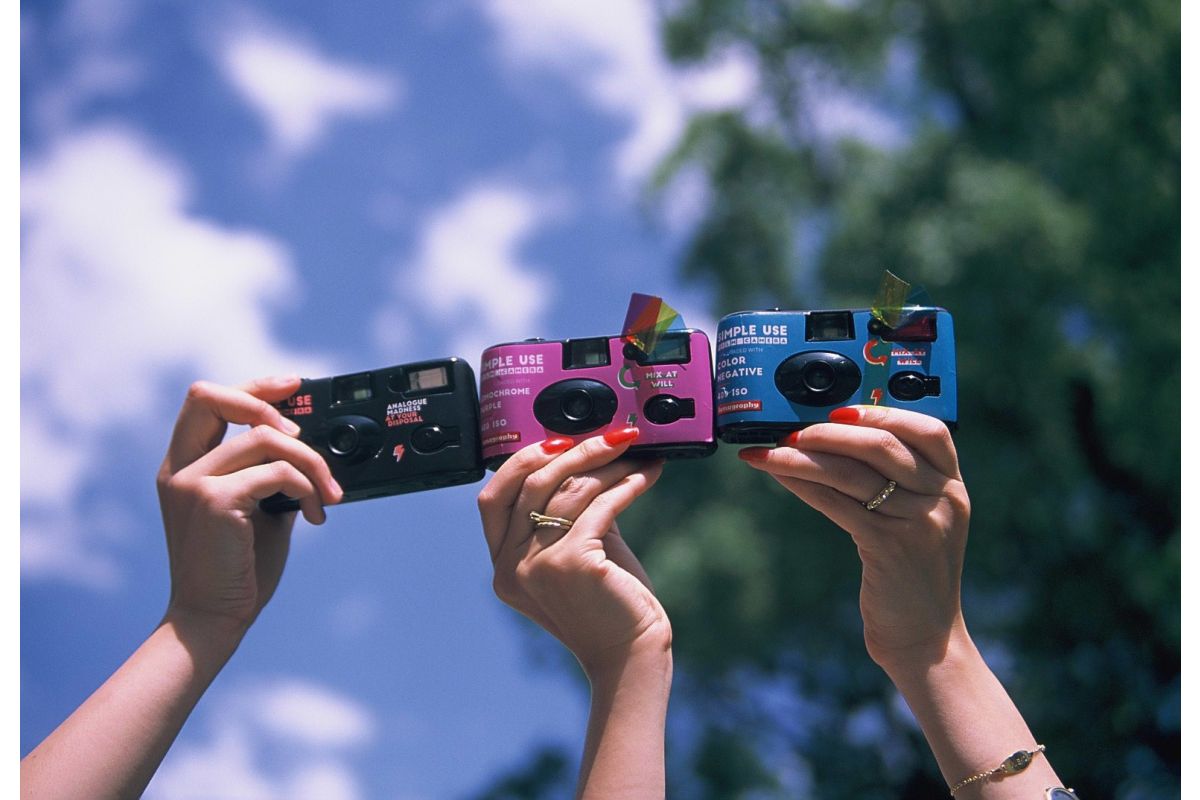 Three retro-style instant cameras in black, pink, and blue are held up by three hands against a bright blue sky with green trees in the background.