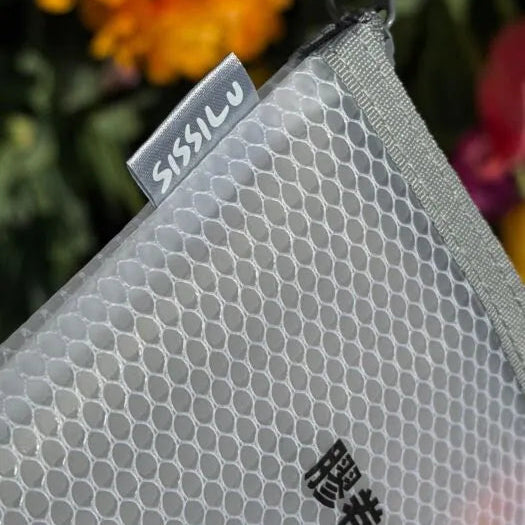 Close-up of a textured gray pouch with 'SissiLu' branding against a blurred floral background.
