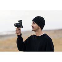 Man with a black beanie holding a black camera with a grip while smiling on a blurry background