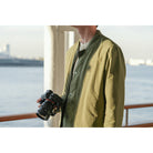 A person wearing an olive green lightweight jacket over a dark shirt, holding a DSLR camera on a balcony with water and a distant city skyline in the background.