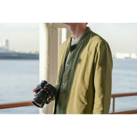 A person wearing an olive green lightweight jacket over a dark shirt, holding a DSLR camera on a balcony with water and a distant city skyline in the background.