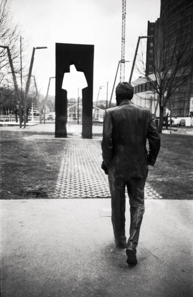 A black and white photograph of a man in a suit walking away from the camera toward a modern abstract metal sculpture in an urban park setting.