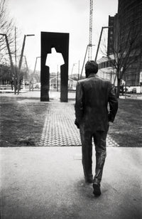 A black and white photograph of a man in a suit walking away from the camera toward a modern abstract metal sculpture in an urban park setting.