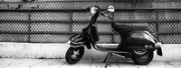 A black vintage-style scooter parked on a sidewalk in front of a chain-link fence, shown in a monochrome photograph.