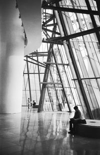 Black and white architectural photograph of a modern glass-walled interior space, with a seated person visible near the lower right and large structural columns and beams throughout the scene.