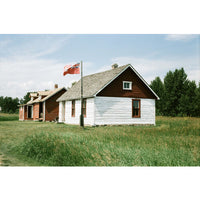 Two older square shaped wood homes, one red and one white and red with a red flag outside and green grasses and trees surrounding