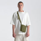 Man wearing a white t-shirt and beige pants with a green crossbody bag on a light gray background