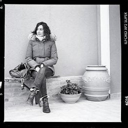 A black and white photograph showing a woman sitting on a stone ledge next to a large ceramic jar, wearing a puffer jacket and jeans. The image has a vintage film border with text reading 'ILIAO VVS SUPER' and '2016'.