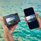 Two hands holding a camera and a smartphone displaying a scenic view of water and sky.